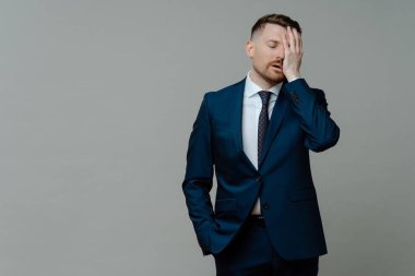 Resmi giyinen genç endişeli adam, iş başarısızlığı konusunda hayal kırıklığına uğramış ve üzgün hissediyor. El ele tutuşup gözlerini kapalı tutarken gri arka planda duruyor. Mesaj için yeri kopyala.