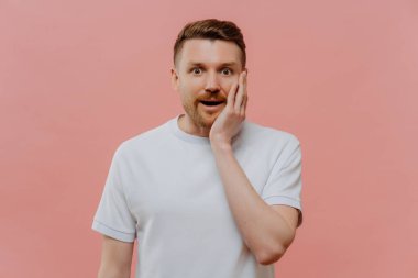 Amazed bearded man keeps hand on cheek stands speechless cannot believe his eyes dressed in casual white t shirt isolated over pink background. Startled unshaven male model finds out shocking news