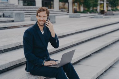 Klasik iki parçalı takım elbiseli gülümseyen iş adamı müşterilerle telefonda konuşuyor. Genç ve çekici iş adamı, merdivenlerde otururken bilgisayarla uzaktan çalışıyor.