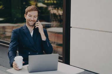 Gülümseyen başarılı iş sahibi uzaktan bilgisayarla çalışıyor ve cep telefonuyla çalışanlarıyla konuşuyor, resmi takım elbise giyiyor, kaldırımdaki kafede oturuyor ve elinde beyaz kahve fincanı tutarak telefon görüşmesi yapıyor.
