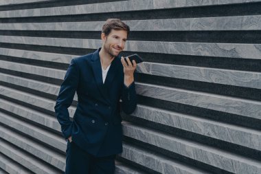 Profesyonel müreffeh sakallı yetişkin adam hoparlör kullanıyor gelen çağrıya cevap veriyor sesli mesaj bırakıyor modern cep telefonunu resmi takım elbise giymiş mermer duvarın yanında duruyor.