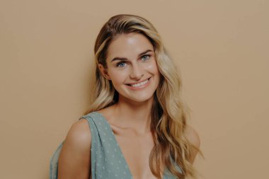 Güzel, narin, mavi elbiseli, parlak tenli genç bir kız kameranın önünde ışıltılı bir gülümsemeyle poz veriyor. Fotoğraf, bej izole arkaplanda, metin için kopya alanı ile birlikte stüdyoda çekildi.