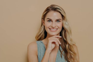 Dalgın, genç, sarışın, mavi elbiseli, çenesinin altından el ele tutuşmuş, gülümseyen ve mutlu bir yüz ifadesiyle kameraya bakan, turuncu arka planda tek başına duran bir kadın.