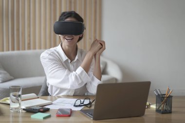 Sanal gerçeklik gözlüklü genç kadın oyuncu, iş yerinde bilgisayarla otururken simülatör oyunu oynuyor. VR kulaklıklı kadın çalışan, yeni teknolojiyi iş yerinde test ediyor.