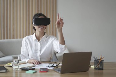VR kulaklıklı kadın ofis çalışanı masa başında otururken dijital arayüzle etkileşime geçiyor, işaret parmağıyla üç boyutlu objelere dokunuyor, mutlu iş kadını sanal gerçeklik gözlüklerini iş dünyası için test ediyor