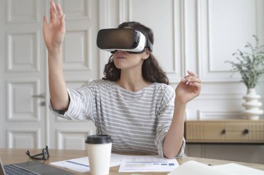 VR kulaklıklı genç kadın banka muhasebecisi ofis masasında oturuyor, dijital arayüzle etkileşimde bulunurken iş için yenilikçi bir cihaz kullanıyor. Siber uzay deneyim kavramı