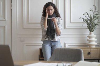 Boynunda sıcak atkısı olan sağlıksız endişeli iş kadını ev ofisinin önünde durmuş termometreye bakıyor termometreye, alnına elle dokunuyor, baş ağrısı ve gripten muzdarip.