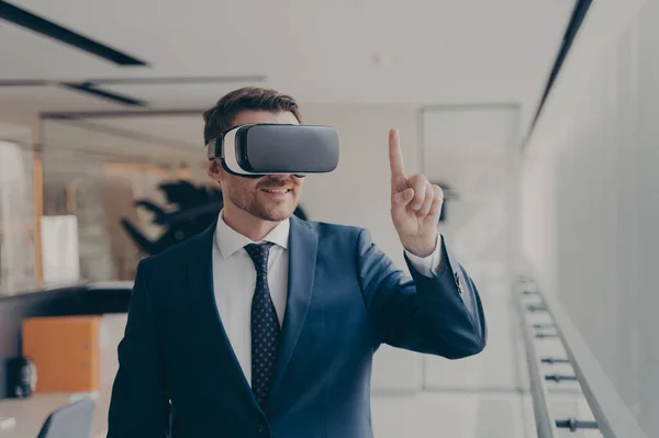 Smiling bearded office employee in suit wearing in VR glasses working in augmented reality in office, businessman in 3d goggles interacting with cyberspace at work, pointing with finger