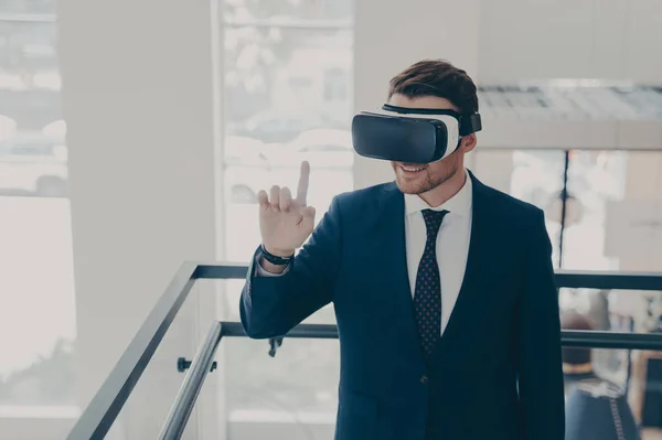 Amazed male office worker in suit being excited while trying out VR glasses, testing virtual reality for business, touching object with forefinger while exploring digital world, standing in office