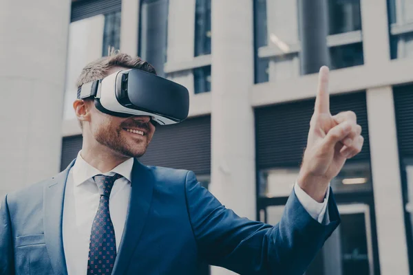 Man with stubble dressed in blue formal suit, wearing VR glasses, viewing something in virtual reality, pointing up with his finger and using it to control, standing next to building