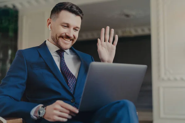 Positive male corporate worker makes video call via laptop computer and earbuds waves hello with palm dressed formally discusses business ideas with partner poses in modern apartment interior