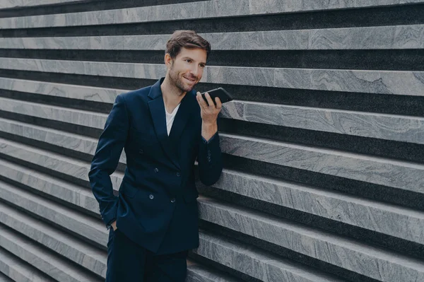 Profesyonel müreffeh sakallı yetişkin adam hoparlör kullanıyor gelen çağrıya cevap veriyor sesli mesaj bırakıyor modern cep telefonunu resmi takım elbise giymiş mermer duvarın yanında duruyor.