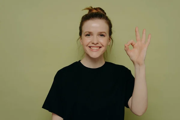 Happy smiling teenage girl with wide smile in black tshirt and hair in bun displaying okay gesture or ok sign with hand, everything going along smoothly, standing in front of green wall