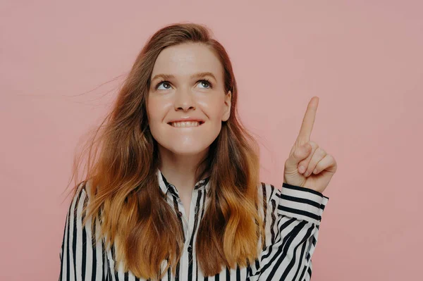 Pretty young woman in formal striped black and white blouse biting lower lip, demonstrating thinking process having pointer finger up posing against pink background. Advertisement and promotion