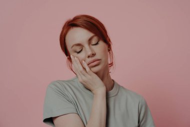 Uykulu, yorgun, kızıl saçlı bir kadın, uyukluyor. Pembe arka planda, kapalı gözlerle, çalışkan bir günün ardından yorgun, kızıl saçlı bir kadının kapalı mekanda çekilmiş görüntüsü.