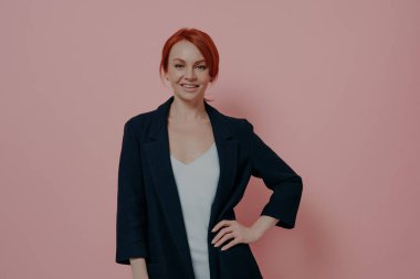 Studio shot of confident beautiful redhead woman in navy jacket smiling at camera and keeping on hand on hip, expressing confidence and success while posing in studio isolated on pink background