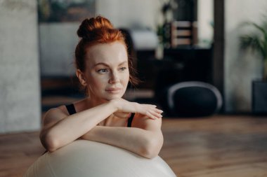 Uzağa bakarken arka planı bulanık olan, fitness stüdyosundaki büyük, beyaz, dengeli spor salonuna dayanan kızıl saçlı genç bir kadının portresi. Spor ve pilates konsepti