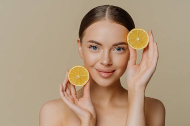 Koyu saçlı esmer kadının portresi doğrudan kameraya bakıyor. İki dilim limon üstsüz duruyor. Bej arka planda sağlıklı, parlak bir cildi var. Yüz bakımı kavramı