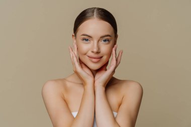 Tatmin olmuş genç bir kadının yüzüne hafifçe dokunduğu, temiz ve sağlıklı bir cilde sahip olduğu banyo havlusuna sarılı minimum makyajlı fotoğrafı, kahverengi arka planda izole edilmiş çıplak omuzları gösteriyor. Güzellik konsepti