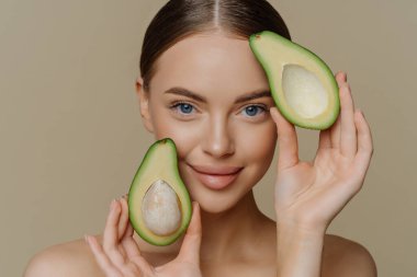Yumuşak mavi gözlü kadının yakın çekim görüntüsü doğrudan kameraya bakıyor. Yüzün yanındaki avokado parçalarına bakıyor. Sağlıklı beslenmeyi seçiyor. güzellik konsepti