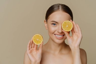 Güzel, narin esmer bir kadının, kameraya nazikçe gülümsemesi, limonun yarısının gözünün üzerinde en sevdiği meyvenin bej arka planda üstsüz poz vermesi. Cilt bakımı kavramı