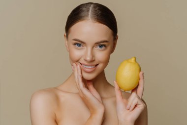 Oldukça sağlıklı Avrupalı bir kadının yatay görüntüsü, doğal yüz maskesi yapmak için limon kullanır. Vücudunun bakımını üstlenir ve kahverengi arka planda kapalı alanda üstsüz kalır. Güzellik konsepti.