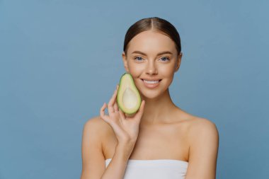 Mutlu esmer kadının yatay pozu Avokado 'nun yarısını yüze yakın tutuyor. Yaşlanma karşıtı organik tarifler kullanıyor. Mavi arka planda izole edilmiş havluya sarılmış olarak kapalı yerde duruyor. Sağlıklı diyet kavramı