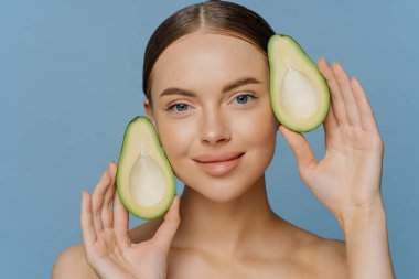 Cilt bakımı için güzellik tedavileri. Memnun esmer, genç Avrupalı kadın model, mavi arka planda izole edilmiş çıplak vücutla yüzünün yanında avokado pozları veriyor. Sağlıklı yağlar. Eko Kozmetik