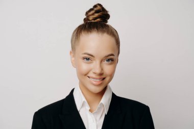 Gülümseyen, koyu renk takım elbiseli, saçları topuz giymiş, geniş gri arka planda sakince ileriye bakan çekici bir iş kadınının portresi.