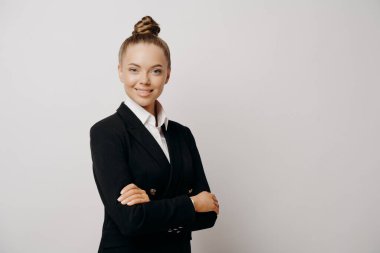Koyu renk takım elbiseli çekici bir kadın yönetici, kollarını çapraz yapıp ileriye bakıyor. Gri arka planda izole edilmiş sakinlik ve kendine güven yayıyor. İş adamları konsepti