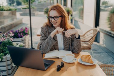 Genç, gülümseyen bir kadın, açık havada kafede oturuyor ve online ders veya dizüstü bilgisayar izliyor, gözlüklü mutlu kızıl saçlı, uzaktan eğitim sırasında bazı notlar alıyor.