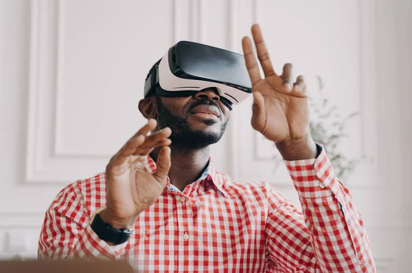 Black man in VR glasses experiencing 3D technology of augmented reality. Excited African male waving with hands up and down makes touching moves with fingers, stands against home office background