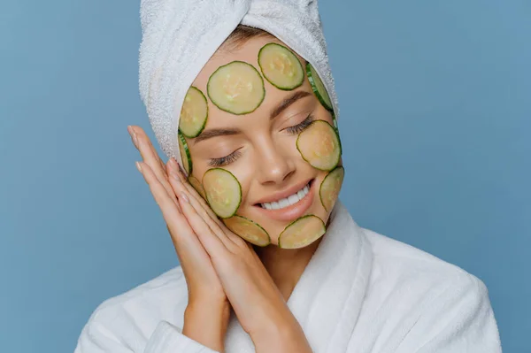 Relaxed satisfied woman tilts head on palms pressed together keeps eyes shut and smiles gently makes facial mask applies cucumber slices on face enjoys morning skin care routine poses indoor