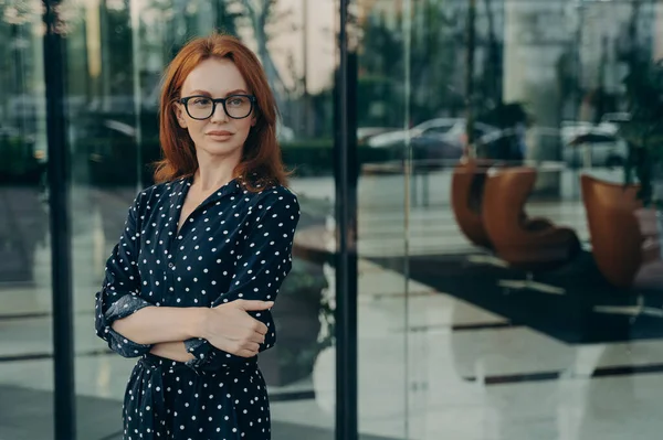 Self assured female director with red hair pleasant appearance dressed in elegant dress spectacles poses near office building waits for partner going to have meeting. Corporate worker outdoor