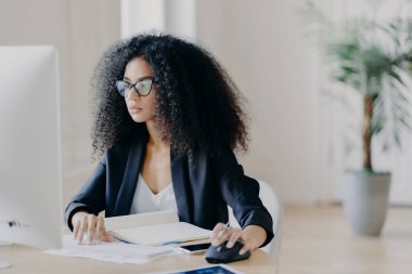 Ciddi kıvırcık iş kadını bilgisayar ekranında çalışıyor, proje üzerinde çalışıyor, ders kitabı ve kağıtlarla çevrili, görüş düzeltmesi için gözlük takıyor, siyah takım elbise, ofiste poz veriyor.