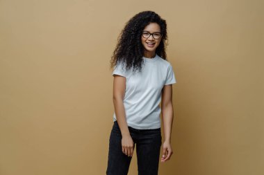 Gülümseyen esmer yüzlü genç bir kadın, optik gözlük takıyor, beyaz tişört ve kot pantolon giyiyor, bej arka planda tasasız poz veriyor. İnsanlar, mutlu ruh hali ve yaşam tarzı konsepti.