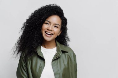 Kaygısız, neşeli, gülümseyen ve kıvırcık saçlı Afro-Amerikan kadın portresi, işte terfi almaktan memnun, mükemmel haberler duyuyor, deri gömlek giymiş, beyaz arka planda soyutlanmış.