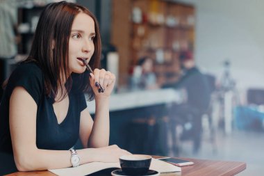 Dalgın kadın blogger blog, yayın için not defteri fikirler yazıyor kafe iç karşı oturur, ağzında kalem tutar, kahve uzakta odaklanmış, giymiş siyah tişörtlü içecekler, planları hakkında düşünüyor