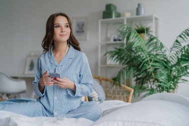 Dalgın kadın modeli çekim film izlemek için akıllı telefon ve kulaklık kullanır, kablosuz internet'e bağlı, çizgili gündelik kıyafeti giymiş, yatakta lotus poz poz veriyor, iyi dinlenme vardır