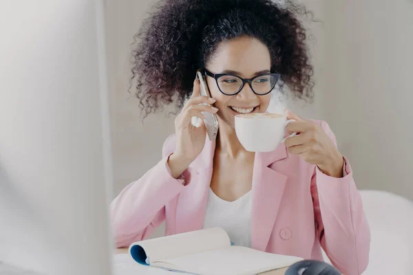 Cropped image of happy young African American woman makes phone call, drinks aromatic latte or espresso, poses in office interior, has pleasant smile wears optical glasses and rosy formal jacket