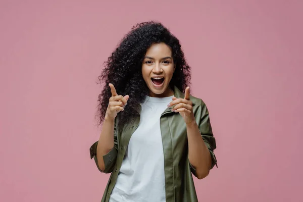 Glad confident woman with Afro hair points index fingers directly at camera, chooses you, picks someone in her team, dressed in white t shirt and leather shirt, poses against pink background