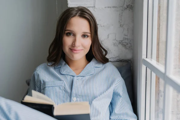 Candid shot of pretty young model sits near window, reads interesting book, looks with satisfied expression at camera, has spare time during weekend. People, leisure, hobby and relaxation concept