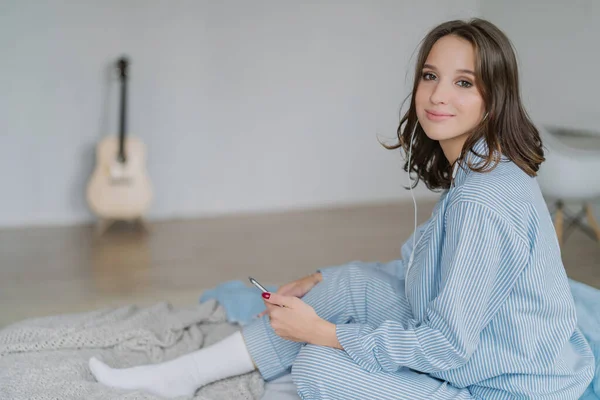 Adorable woman listens lecture on news in internet via cell phone and earphones, has recreation time, connected to wifi at home, poses in bedroom, blurred background with guitar in background