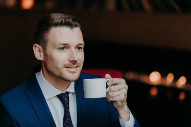 Thoughtful handsome young businessman in formal suit, holds cup of coffee, being deep in thoughts, thinks about new startup, recreats during dinner break at restaurant. People, business concept