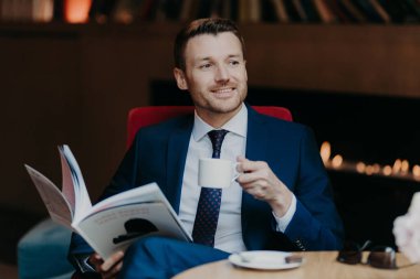 Cheerful unshaven male manager or employee has pleased expression, holds cup of coffee, rests in cafeteria, holds popular magazine, being focused aside, notices his colleague, dressed formally