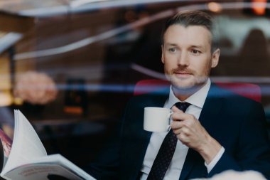 View from window of handsome successful young male boss, holds cup with hot aromatic beverage, looks through menu in luxury restaurant, dressed in formal expensive suit, thinks about something