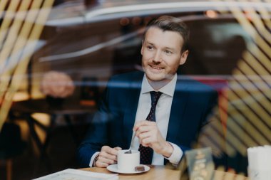 Optimistic cheerful young businessman has thoughful expression, dressed in formal clothes, looks through window in cafeteria, satisfied with results of work. People, leisure and business concept