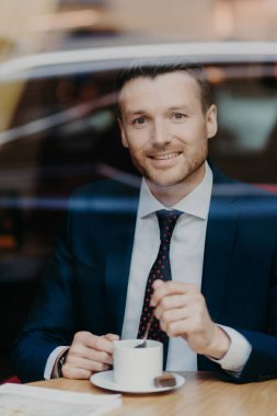 Verrtical shot of delighted handsome male professional worker in black suit, drinks aromatic beverage, looks joyfully at camera, poses in restaurant, sits near window, has coffee break after work