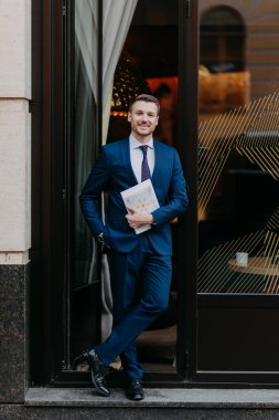 Vertical shot of successful male executive worker in formal suit, keeps hand in pocket, keeps legs crossed, holds journal, stands near cafe, has positive expression. Business and work concept