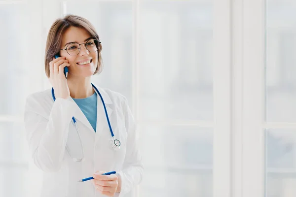 Shot of pleased female physician or surgeon gives prescription via mobile phone, has conversation with patient, holds pen, wears white uniform, works in hospital, stands indoor against window